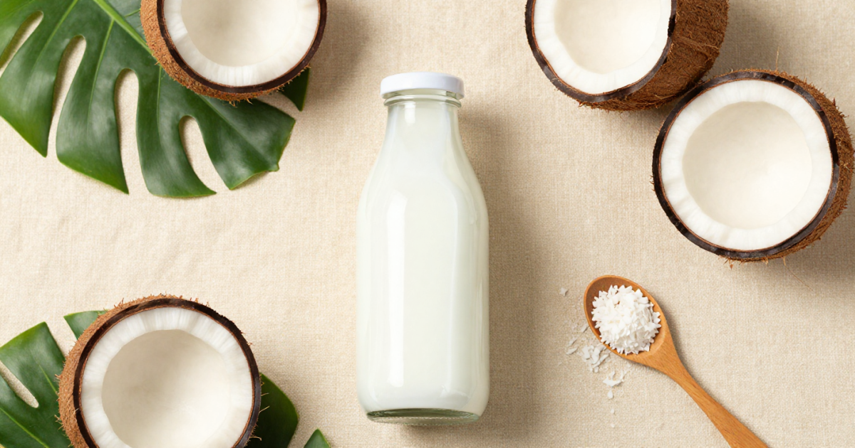 Botella de leche de coco rodeada de mitades de coco, hojas tropicales y una cuchara con coco rallado sobre fondo beige, representando el uso cotidiano y natural de la leche de coco.