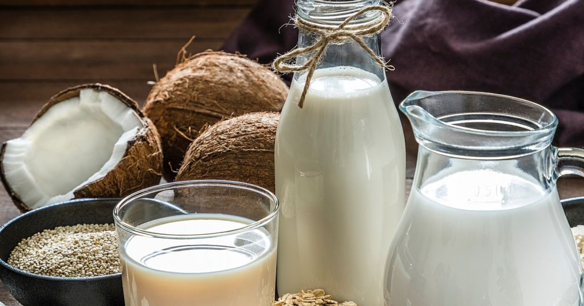 Botellas y vasos de leche vegetal junto a cocos, avena y granos sobre una mesa de madera, representando la comparación entre leche de coco, almendra, soya y avena.